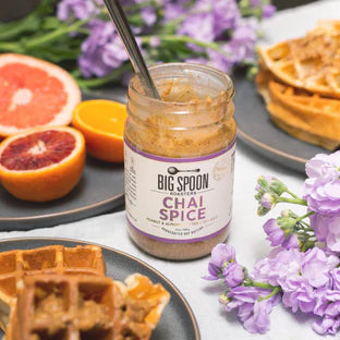 A jar of Chai Spice Peanut & Almond Butter is open on a table with purple flowers, waffles, and halved citrus fruits