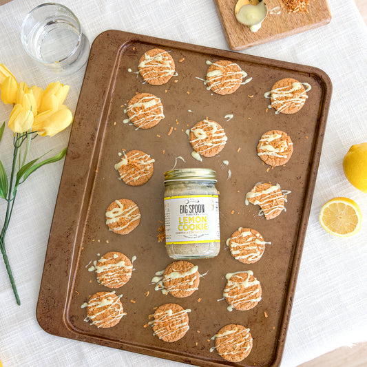 A jar of Lemon Cookie Coconut & Cashew Butter lies on a cookie sheet with lemon cookies surrounding it