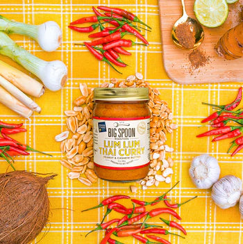 13oz glass jar of Lum Lum Thai Curry nut butter on golden fabric surrounded by chilis, lemongrass, coconut, peanuts, cashews, and shallots.