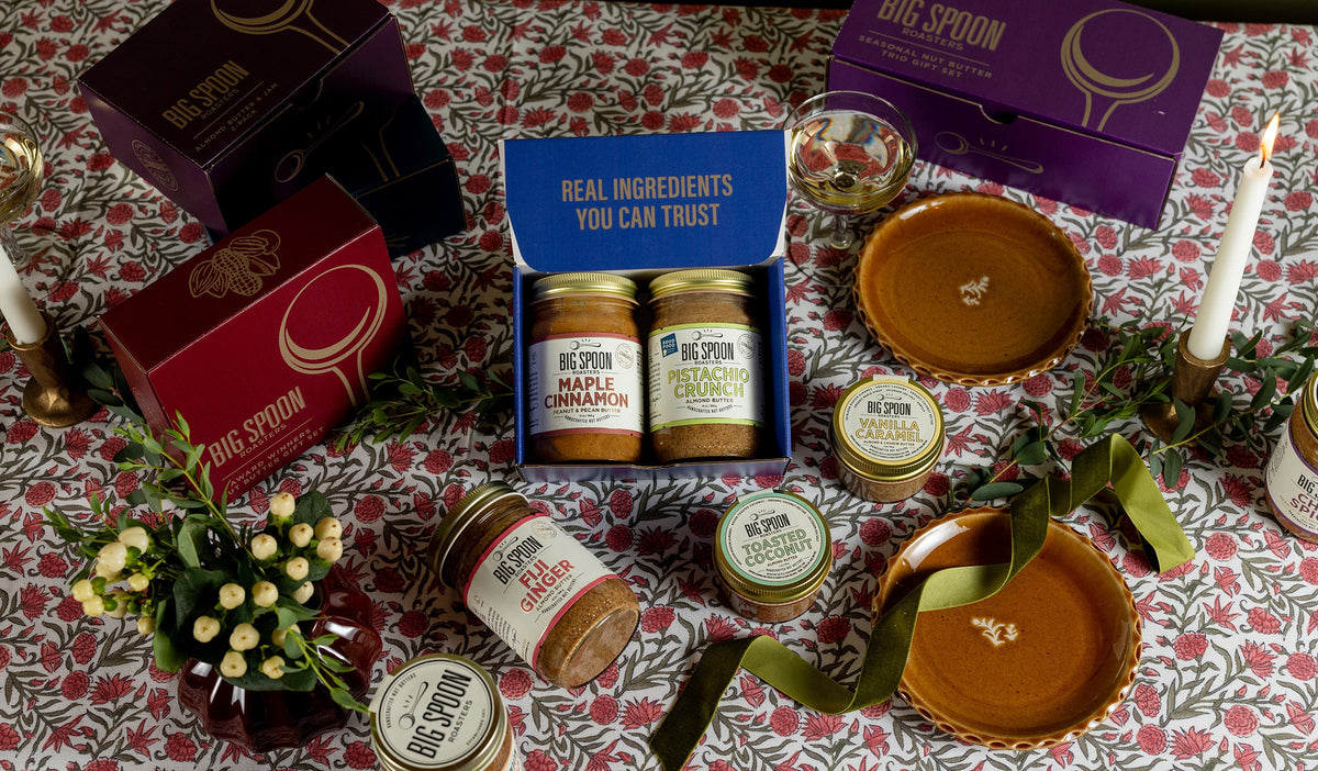 A hand reaches for a 3oz jar of nut butter surrounded with other nut buttes and sliced bread all on a white knit picnic blanket