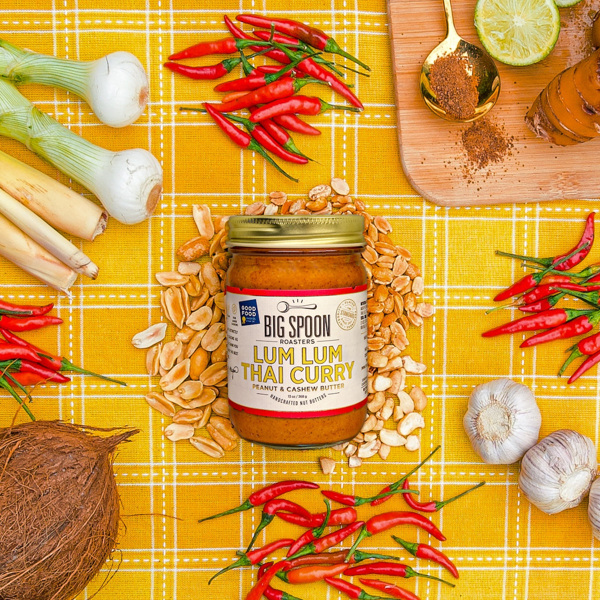 A 13oz jar of Lum Lum Thai Curry nut butter is surrounded by peanuts cashews, red chilies, shallots, coconut, lime, and ginger all on a yellow gingham cloth