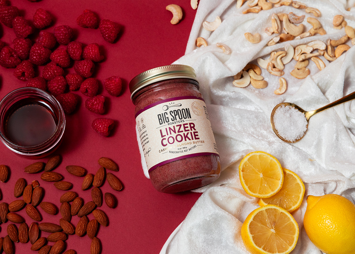 Linzer Cookie Cashew & Almond Butter surrounded by raspberries, maple syrup, almonds, cashews, sea salt, and lemon on a split red and white velvet background