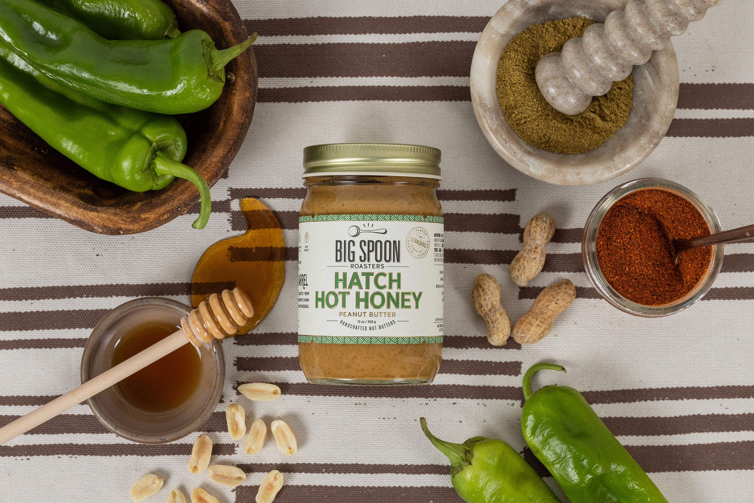Jar of Hatch Hot Honey Peanut Butter surrounded by peanuts, hatch chiles, and honey