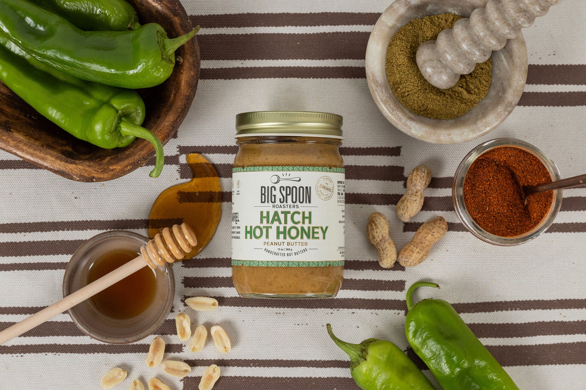 Jar of Hatch Hot Honey Peanut Butter surrounded by peanuts, hatch chiles, and honey