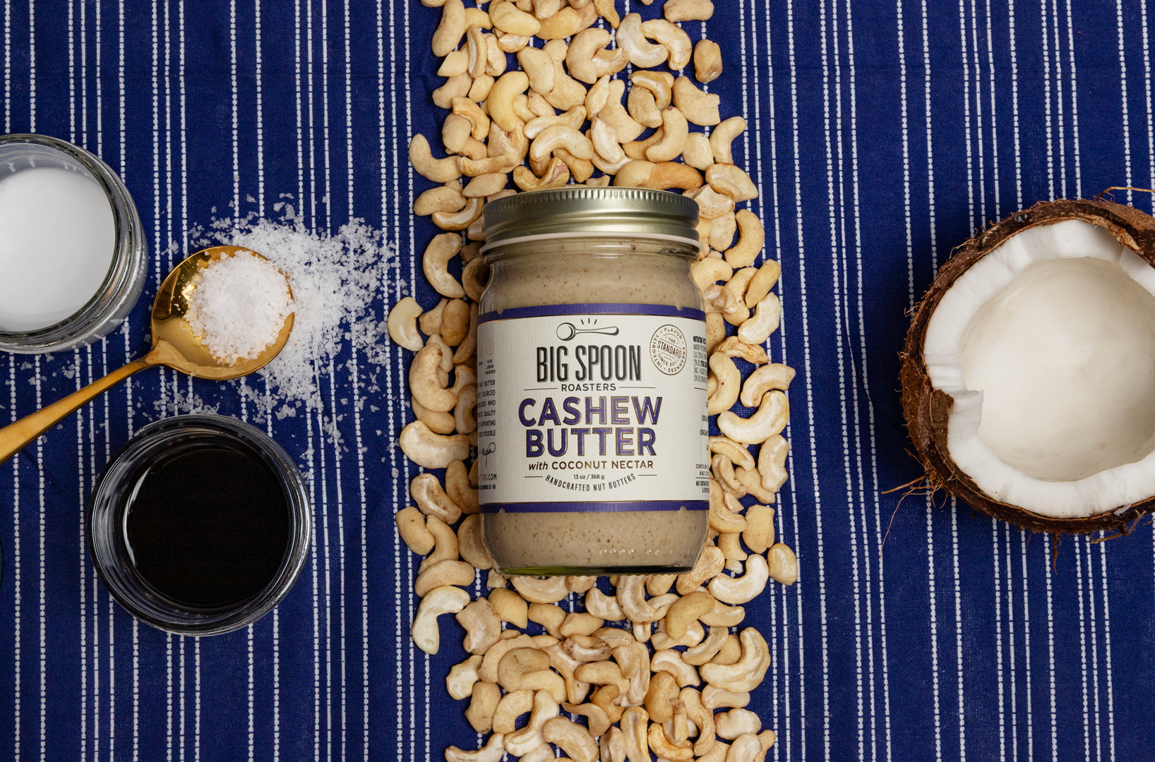 Jar of Big Spoon Cashew Butter surrounded by cashews, coconut, coconut nectar and oil, and salt on a blue striped background