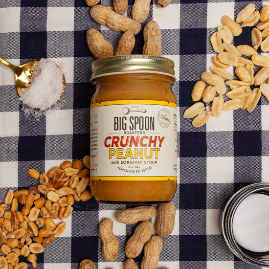 Jar of Big Spoon Crunchy Peanut on a checkered tablecloth with peanuts, coconut oil, sorghum, and salt around