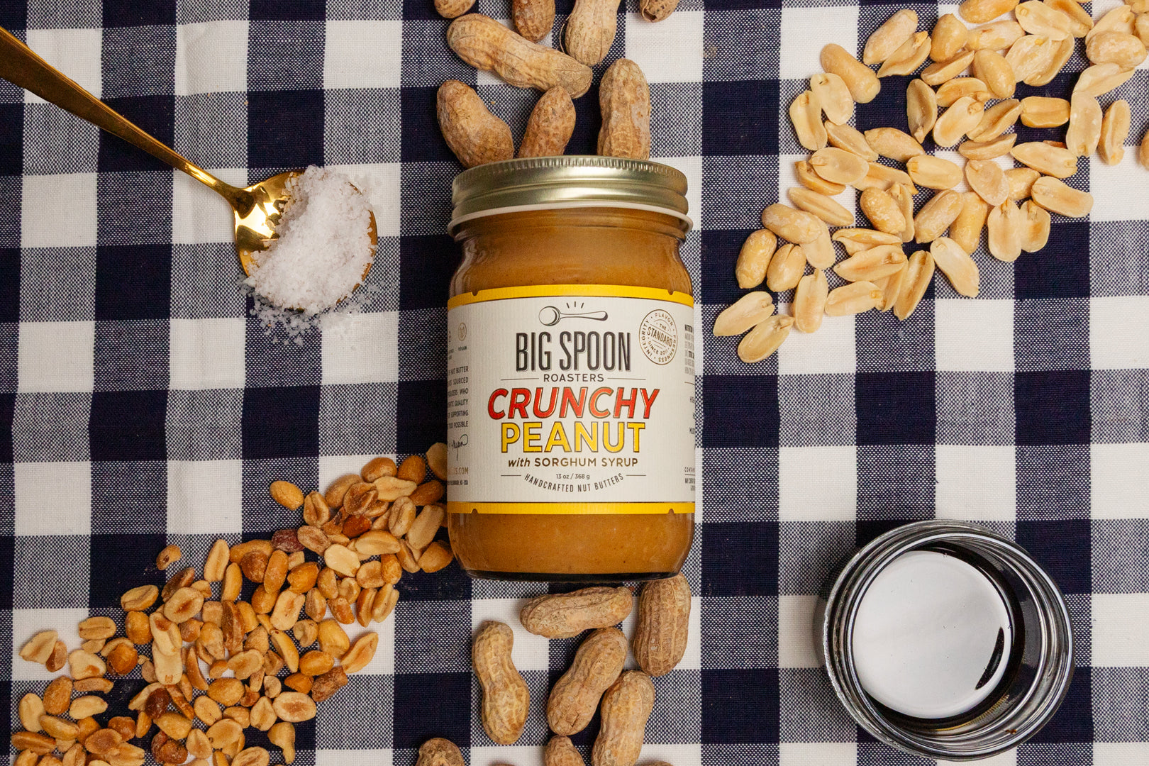 Jar of Big Spoon Crunchy Peanut on a checkered tablecloth with peanuts, coconut oil, sorghum, and salt around