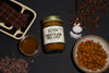 a jar of Chocolate Sea Salt almond butter surrounded by a dish of chocolate bars, a jar of honey, a spoonful of sea salt, chocolate chips, almonds, and cacao nibs