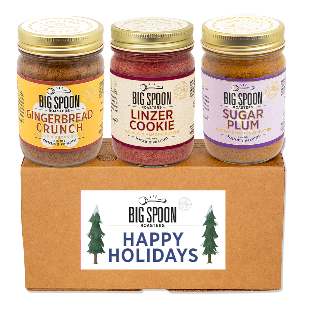 Three jars of Big Spoon Roasters holiday-themed nut butters - Gingerbread Crunch, Linzer Cookie, and Sugar Plum - on a cardboard box with a 'Happy Holidays' label.