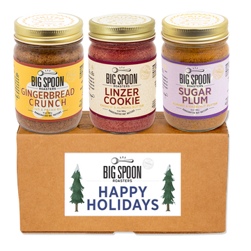 Three jars of Big Spoon Roasters holiday-themed nut butters - Gingerbread Crunch, Linzer Cookie, and Sugar Plum - on a cardboard box with a 'Happy Holidays' label.
