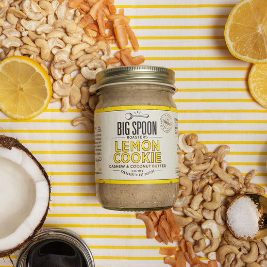 Jar of Lemon Cookie Cashew Butter with lemon, coconut, cashews, salt, and coconut nectar/oil around it on a yellow striped background
