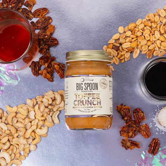 Jar of Big Spoon Toffee Crunch surrounded by cashews, pecans, peanuts, salt, and small bowls of syrup on a metallic background.