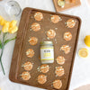 13oz jar of Lemon Cookie Cashew Butter on a cookie sheet surrounded by lemon cookies on a light linen cloth table surrounded by a fresh cut lemon, a cutting board, a glass of water, and a small bunch of yellow tulips.