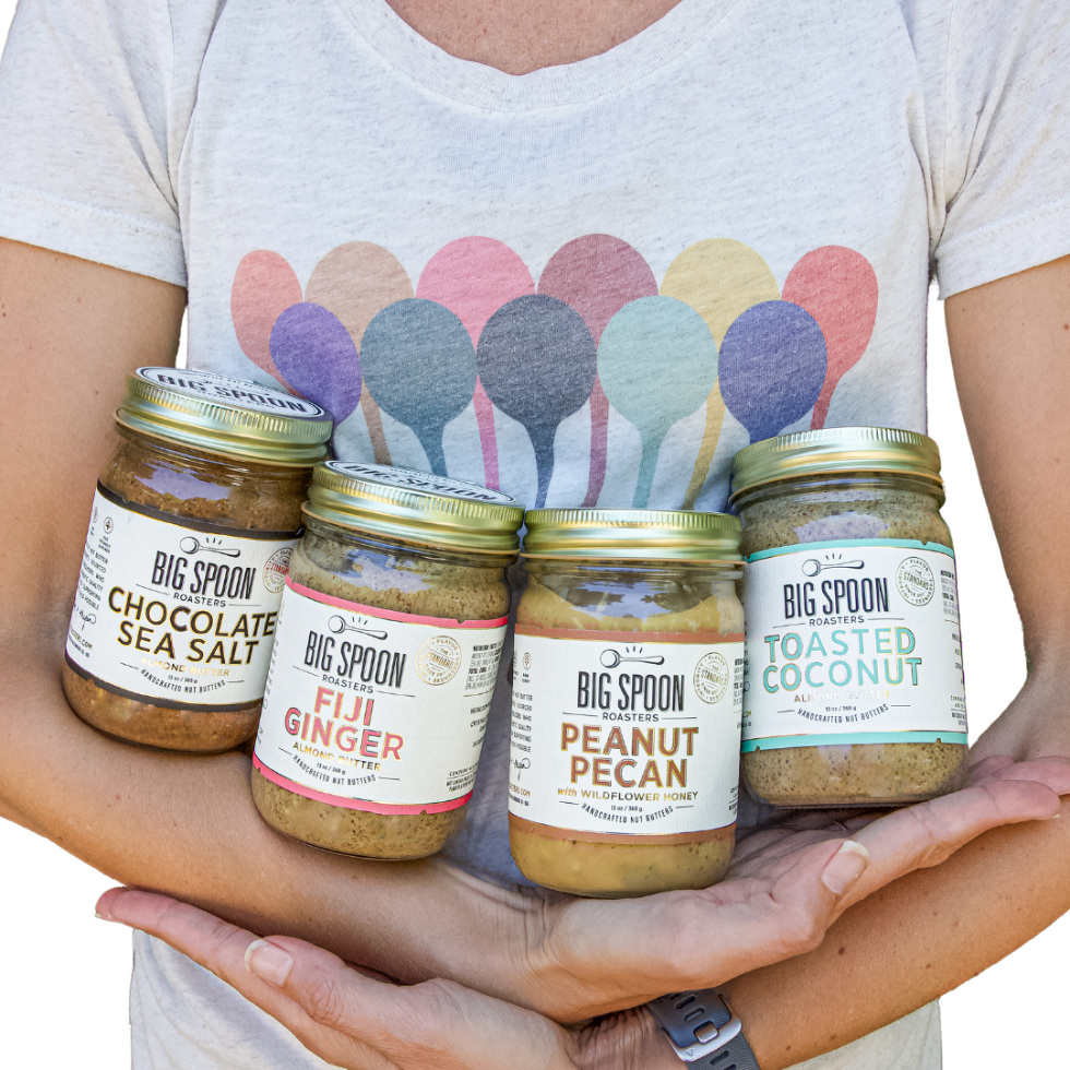 A person in a colorful spoon shirt holds four jars of Big Spoon Roasters nut butters