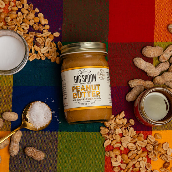 Jar of Big Spoon Peanut Butter surrounded by peanuts, coconut oil, honey, and salt on a colorful checkered background