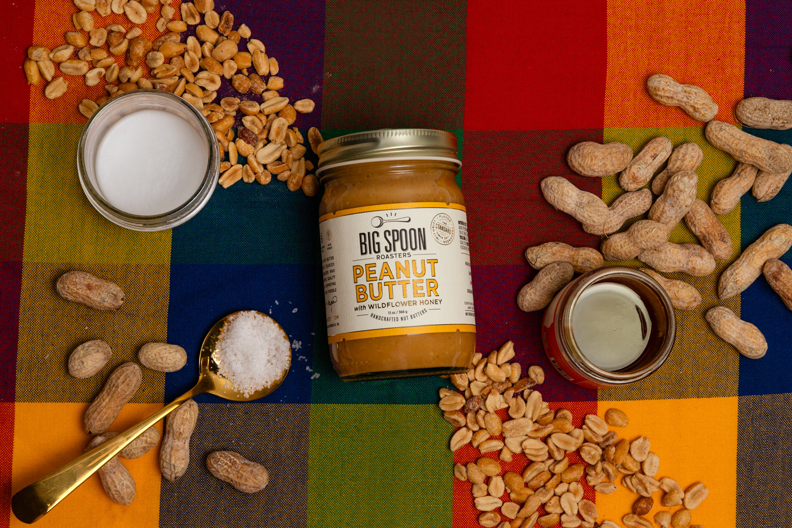 Jar of Big Spoon Peanut Butter surrounded by peanuts, coconut oil, honey, and salt on a colorful checkered background