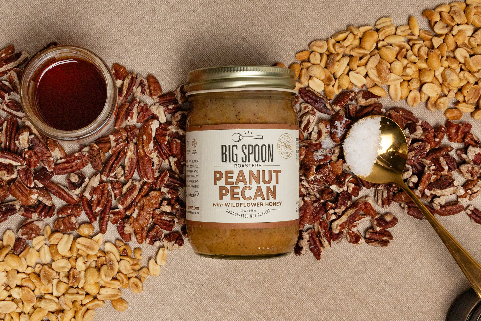 Jar of Big Spoon Roasters Peanut Pecan spread with ingredients and a spoon on a beige background
