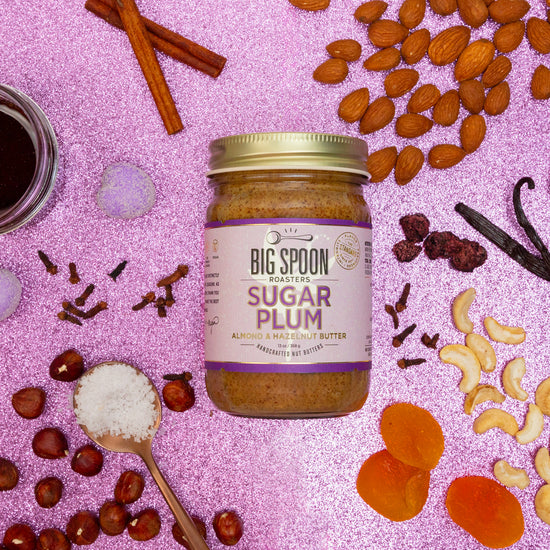 Photo of Sugar Plum Almond & Hazelnut Butter  surrounded by almonds, cashews, hazelnuts, maple syrup, cloves, cranberries, apricots, vanilla, and cinnamon on a glittery pink-purple background