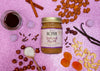 Photo of Sugar Plum Almond & Hazelnut Butter  surrounded by almonds, cashews, hazelnuts, maple syrup, cloves, cranberries, apricots, vanilla, and cinnamon on a glittery pink-purple background