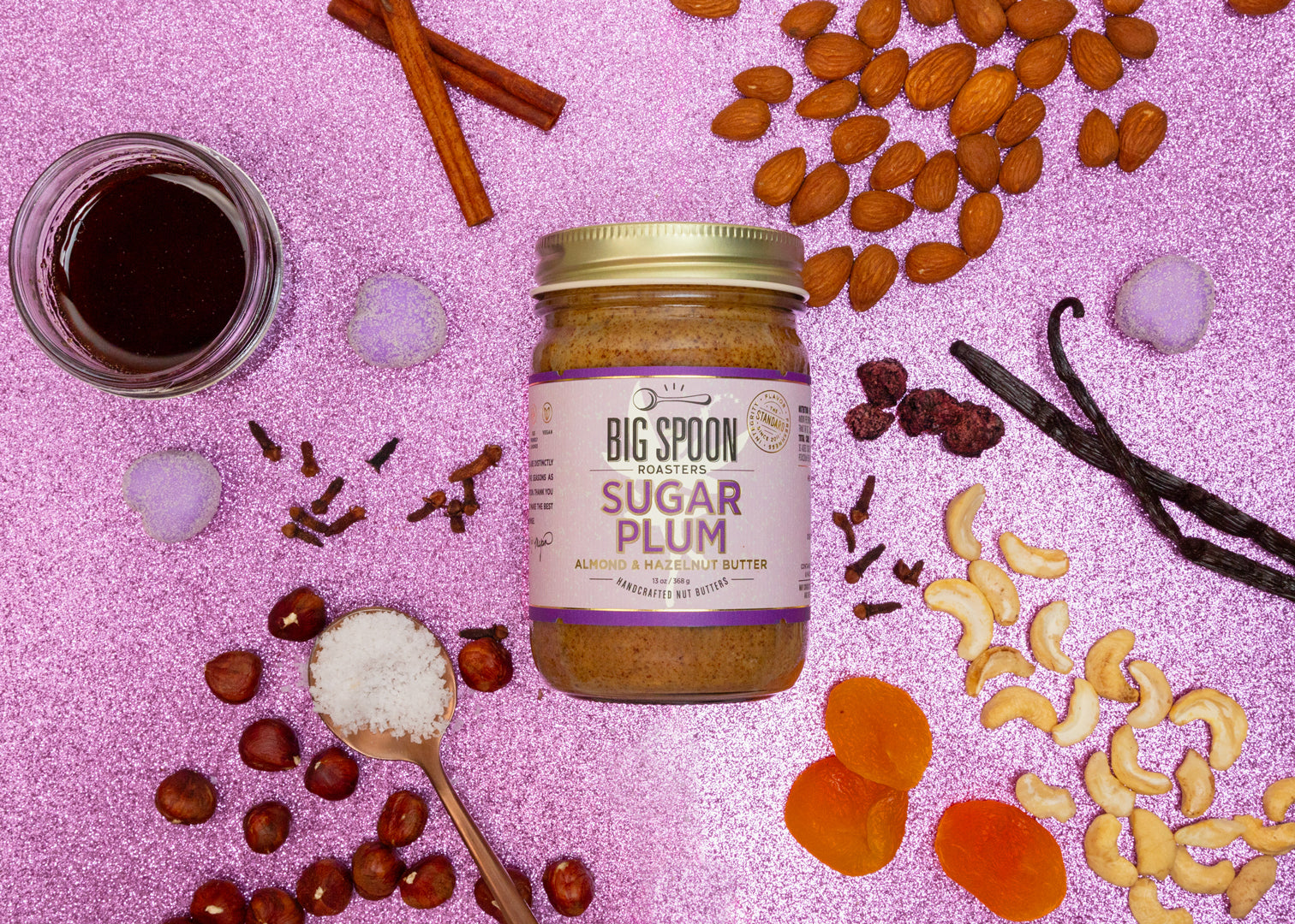 Photo of Sugar Plum Almond & Hazelnut Butter  surrounded by almonds, cashews, hazelnuts, maple syrup, cloves, cranberries, apricots, vanilla, and cinnamon on a glittery pink-purple background