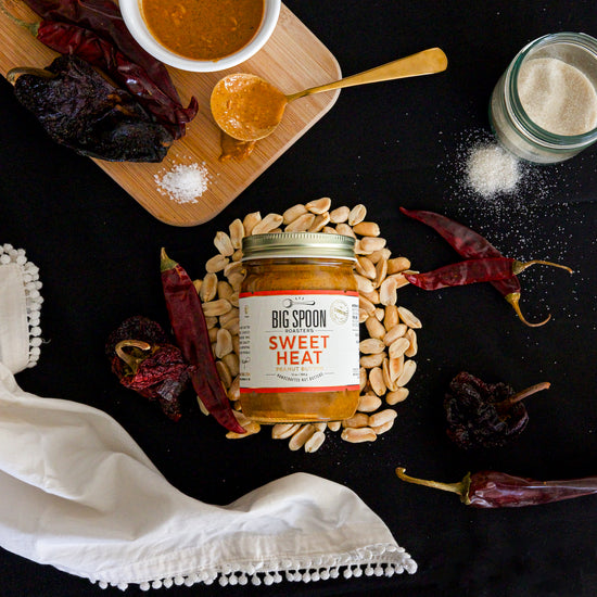 13oz jar of Sweet Heat Peanut Butter on a black table cloth surrounded by peanuts, chilis, sugar, and a golden spoon with Sweet Heat Peanut Butter on it