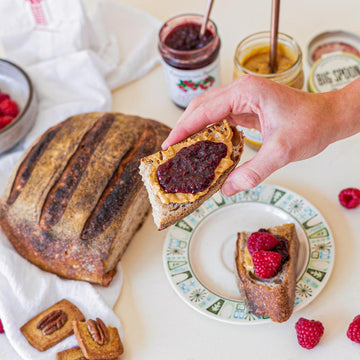 A hand holds a piece of peanut butter and jelly toast in front of a spread of bread, peanut butter, jam, and fresh raspberries