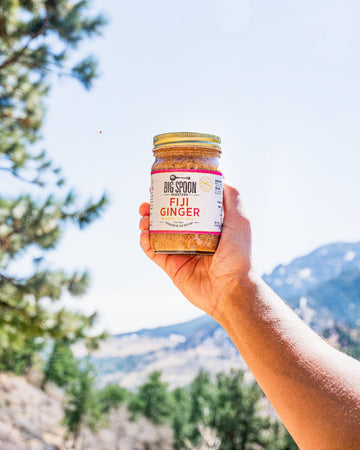 A hand holds a jar of Fiji Ginger in front of a mountain landscape