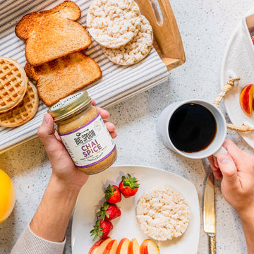 Hand holds a jar of Chai Spice nut butter while the other hand holds a cup of coffee, surrounded by toast, waffles, rice cakes, and fresh fruit