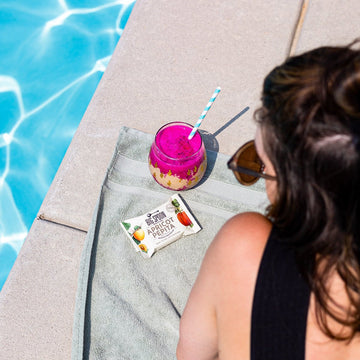 A woman looks over her shoulders wearing sunglasses and looking at an apricot pepita nut butter bar beside a glistening pool