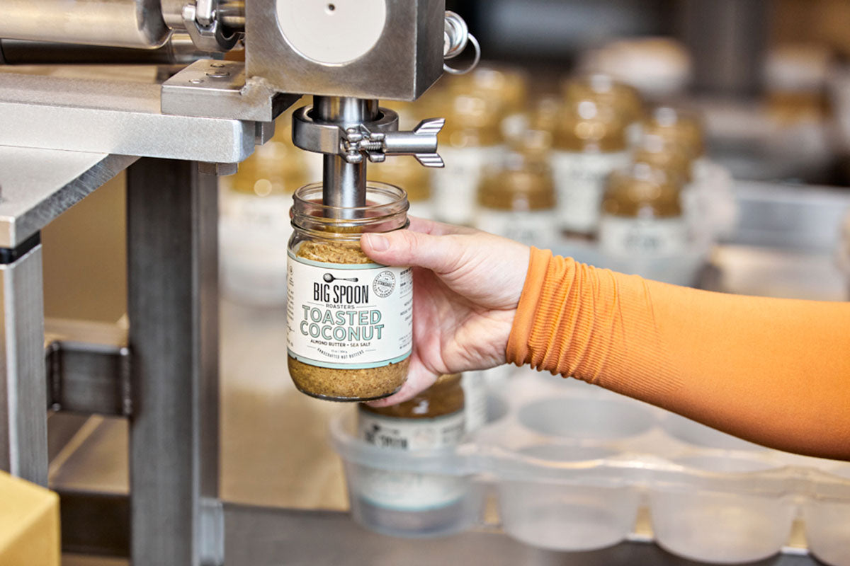 A hand holding a jar of Big Spoon Roasters nut butter under a filling machine.